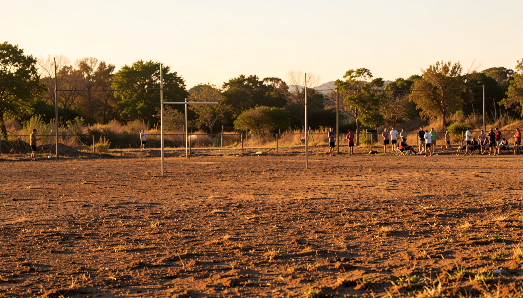 Dusty outdoor sports field in a rural setting, warm golden-hour light, simple goalposts and a gathering of spectators in the distance, earthy browns and greens