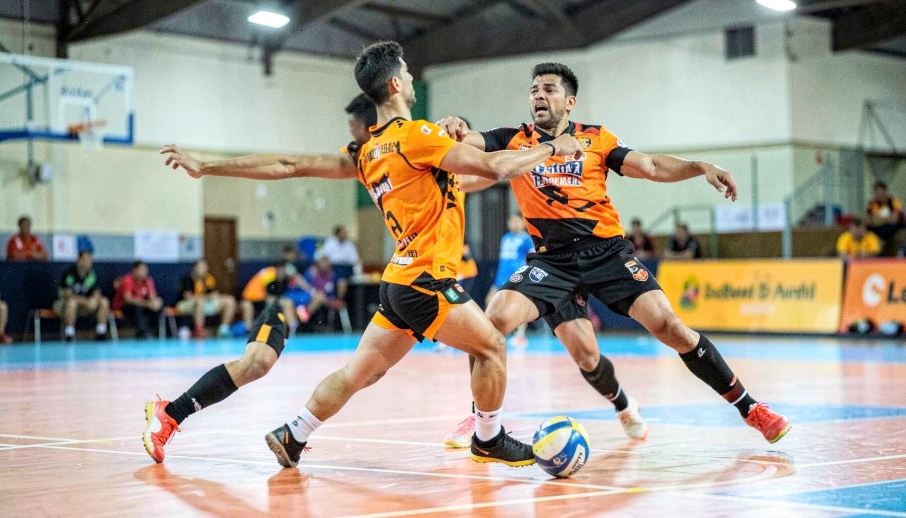 Kabaddi player in Bengaluru Bulls orange and black kit, mid-raid on a brightly lit indoor court