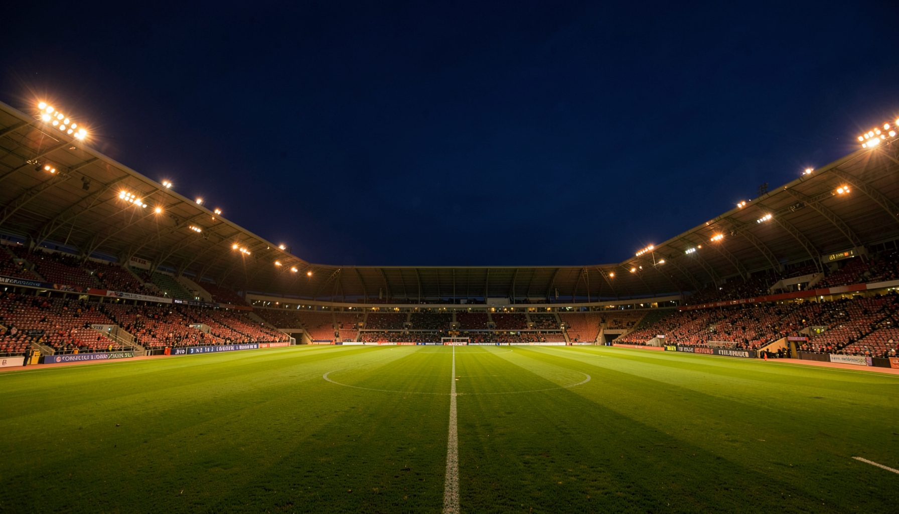 Wide-angle shot of a floodlit sports stadium at dusk, deep navy sky with warm amber field lights, empty green pitch ready for action