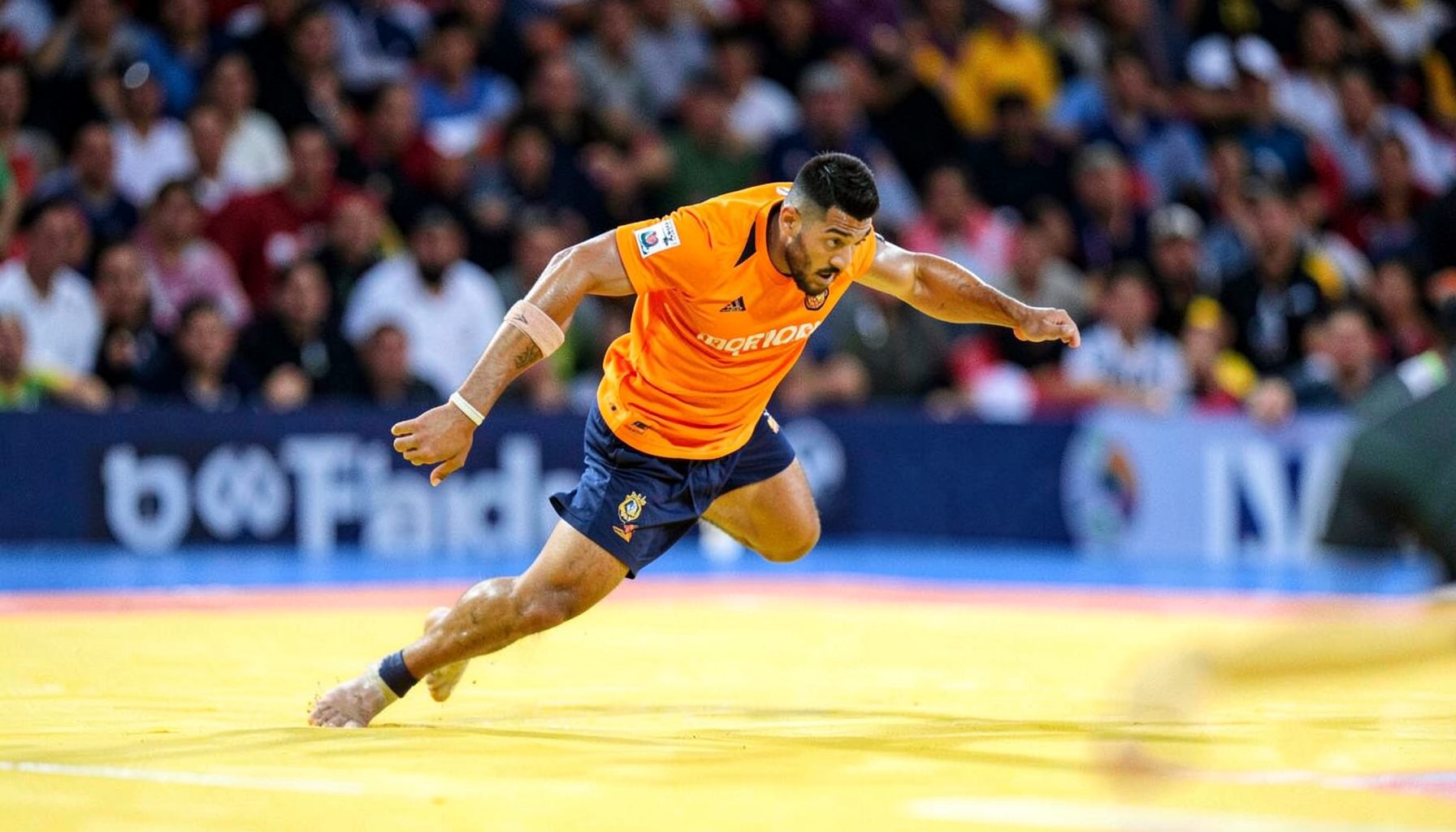 Kabaddi raider in bright orange jersey lunging into opponent territory on a yellow mat, crowd blurred behind