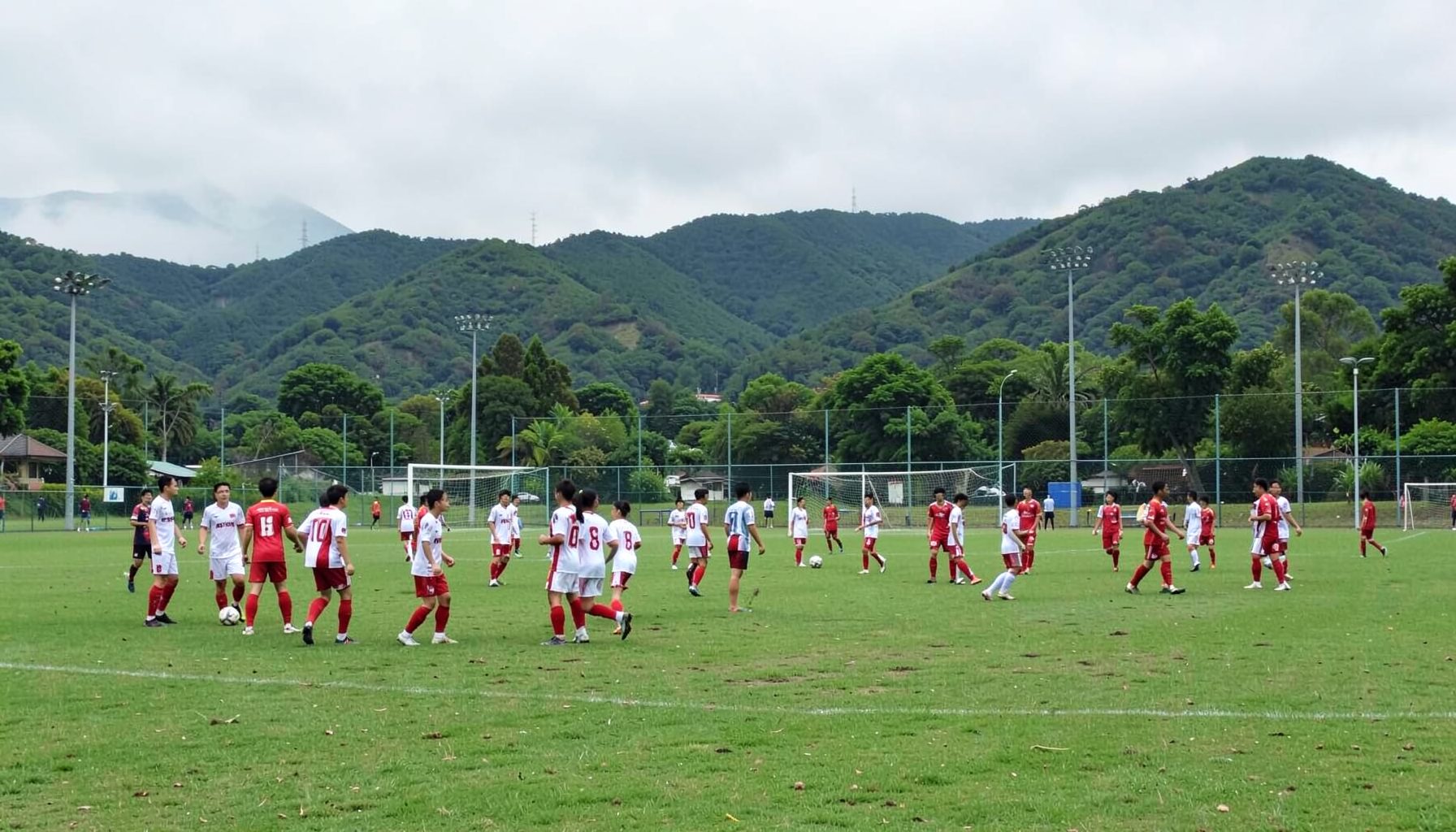 Outdoor community sports field with teams in white and red kits, green hills in the background under overcast sky